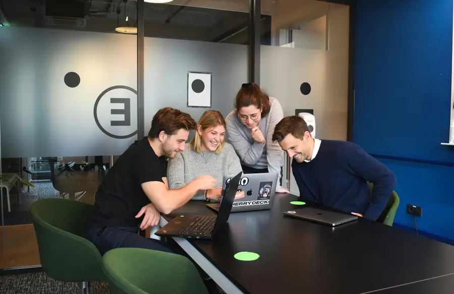 Four colleagues laughing together around a laptop at a meeting table